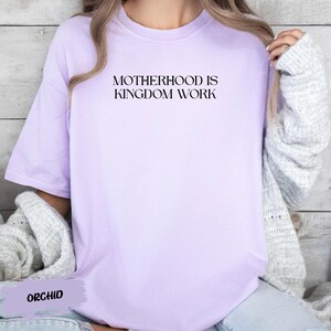 A person wearing a purple t-shirt with the text "MOTHERHOOD IS KINGDOM WORK" printed on it.