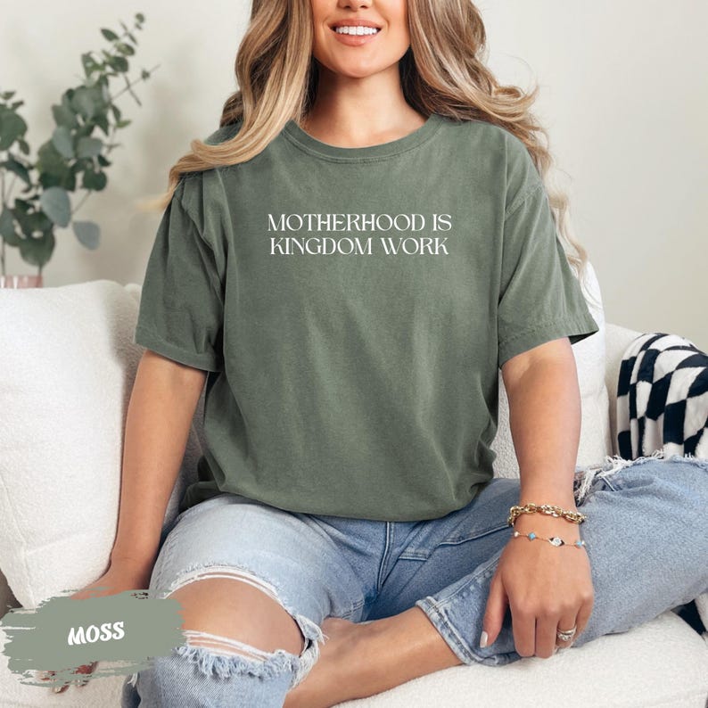 A woman wearing a green t-shirt with the text "MOTHERHOOD IS KINGDOM WORK" is sitting on a couch, smiling.