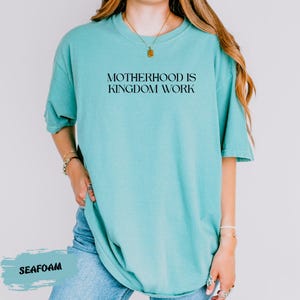 A woman wearing a turquoise t-shirt with the text "MOTHERHOOD IS KINGDOM WORK" printed on it.