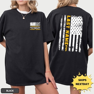 May include: A black t-shirt featuring a distressed American flag graphic on the front left chest with the text YOUR NAME. The back displays a large, vertical distressed American flag graphic with the text LAST NAME in yellow.