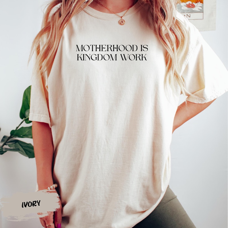 A person wearing a white t-shirt with the text "MOTHERHOOD IS KINGDOM WORK" printed on it, standing in front of a plant.