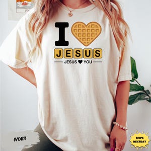 A person wearing a white t-shirt with a graphic design featuring a heart-shaped waffle and the text "I ♥ JESUS" and "JESUS HEARTS YOU".
