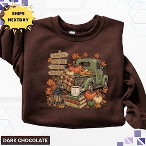May include: A dark chocolate brown crewneck sweatshirt featuring a graphic print of a vintage green pickup truck loaded with pumpkins, a plaid blanket, a lantern, and a stack of books with a mug labeled COZY SEASON and a sign reading PUMPKINS.