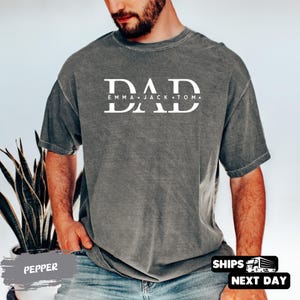 A man wearing a gray t-shirt with the text "DAD" printed on it, standing in front of a plant.