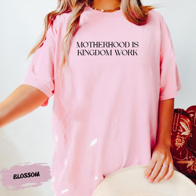 A person wearing a pink t-shirt with the text "MOTHERHOOD IS KINGDOM WORK" printed on it.