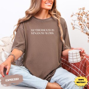 A woman wearing a brown t-shirt with the text "MOTHERHOOD IS KINGDOM WORK" is sitting on a couch, holding a coffee mug.