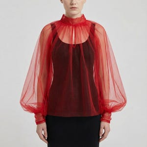 Puede incluir: Una blusa roja transparente con cuello alto y mangas largas y abullonadas. La blusa se superpone a una parte superior oscura y se combina con una falda negra. La tela parece ser tul ligero, creando un aspecto delicado y aireado.