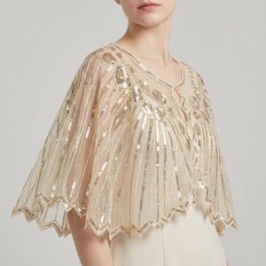 May include: A beige capelet, embellished with gold sequins. The capelet has a scalloped hem and detailed sequin patterns, creating a shimmering effect. It features a keyhole neckline and a delicate, flowing silhouette, suitable for formal occasions.