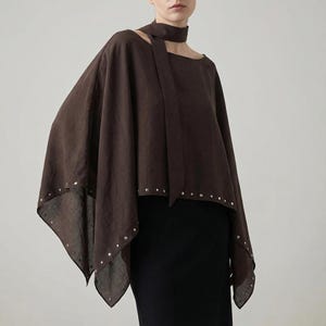 May include: A brown, draped top with a unique asymmetrical design. The top features a high neckline with a scarf detail and is adorned with small silver studs along the edges. The garment is paired with a black skirt, creating a minimalist, modern aesthetic.