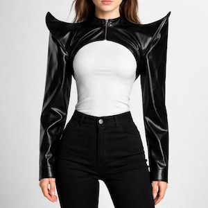 Puede incluir: Chaqueta bolero de cuero sintético negro con detalles de hombros puntiagudos y dramáticos. La chaqueta tiene mangas largas y cremallera frontal. La modelo lleva un top blanco y pantalones negros.