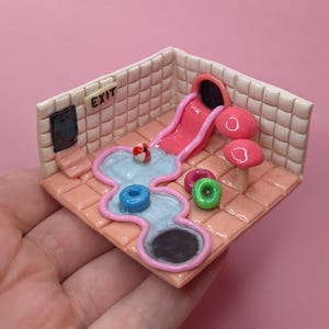 Könnte beinhalten: Ein Miniatur-Diorama mit einer rosa Rutsche, einem Pool und Pilzen. Die Szene umfasst einen blauen Donut, einen rot-weißen Ball und einen grünen Donut. Die Wände sind gefliest, und das Wort "EXIT" ist sichtbar.
