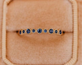 May include: A gold band ring featuring a series of nine round, bezel-set blue sapphires arranged in a horizontal row. The metal has a textured, matte finish, and the stones vary slightly in size, creating a delicate and consistent line across the finger.