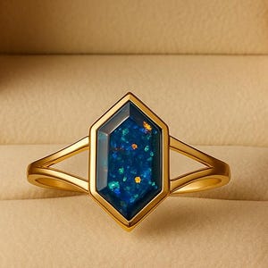 May include: A gold ring with a blue gemstone in a hexagonal shape, set in a gold frame. The ring is displayed in a beige jewelry box with the brand name "RIOLUXEUS" in gold lettering.