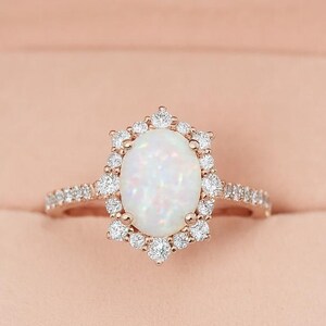 Peut inclure: Une bague en or rose avec une grande pierre centrale ovale en opale, entourée d'un halo de petites pierres précieuses transparentes. La bande est également ornée de petites pierres précieuses transparentes. La bague est présentée dans un écrin en velours rose.