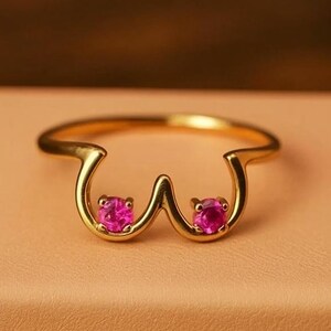 May include: A gold-colored ring with a unique design featuring two pink gemstones. The ring is set on a light brown box with the word "RIOLUXEUS" printed on the front. The ring's design is a stylized representation of a pair of breasts.