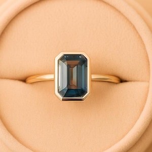May include: A gold ring featuring an emerald-cut, teal gemstone. The ring is set in a gold band and presented in a peach-coloured ring box. The gemstone has a rectangular shape with a step-cut design.
