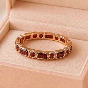 Op de afbeelding: Een goudkleurige ring met rechthoekige en ronde rode edelstenen, gepresenteerd in een roze fluwelen sieradendoos. De doos is bedrukt met de tekst "Rioluxeus".
