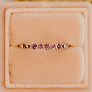 May include: A gold ring with a row of small, round, pink gemstones. The ring is displayed in a pink velvet ring box, with a gold-colored frame in the background. The ring is a delicate piece of jewelry.