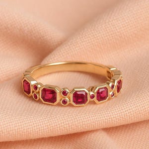 May include: A gold ring featuring alternating square and round red gemstones. The ring is displayed on peach-coloured fabric. The gemstones are evenly spaced around the band, creating a continuous pattern. The ring's design is elegant and eye-catching.