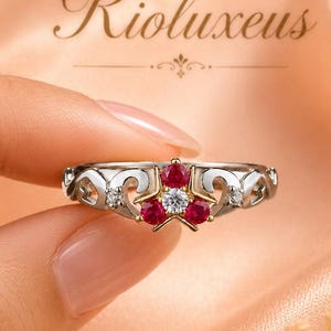 Könnte beinhalten: Ein silberfarbener Ring mit einem sternförmigen Mittelstück aus drei runden roten Steinen und einem klaren Stein in der Mitte. Die Ringschiene weist filigrane Ausschnitte und kleine klare Steine an den Seiten auf. Der Text Rioluxeus ist sichtbar.