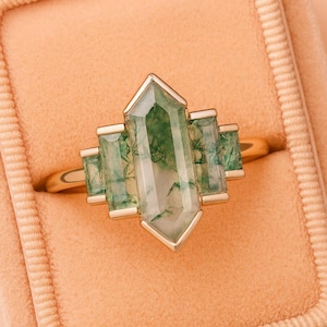 Puede incluir: Un anillo de oro con una piedra preciosa rectangular de ágata musgosa verde y un diseño geométrico. El anillo tiene una banda de oro y se presenta en un joyero de color melocotón.