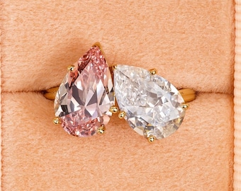 May include: Two pear-shaped diamond rings set in gold, one pink and one clear, displayed in a pink velvet ring box. The rings are side-by-side, showcasing their brilliance and intricate facets.
