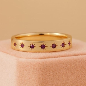 Peut inclure: Une bague dorée ornée d'une série d'étoiles, chacune sertie d'une petite pierre rouge foncé. La bague est posée sur une surface rose, avec un fond neutre. La bague est de style jonc, avec une finition lisse et polie. Le design est symétrique.