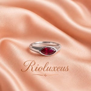 Op de afbeelding: Een zilveren ring met een marquise-geslepen robijn. De ring ligt op een perzikroze satijnen stof. Het woord "Rioluxeus" staat in een sierlijk lettertype op de stof.