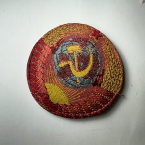 May include: A round, embroidered patch with a red, yellow, and teal design. The central image features a hammer and sickle, surrounded by a wheat wreath. The patch has a textured appearance and a stitched border.