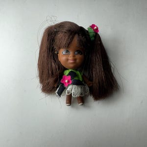 May include: A small doll with brown hair styled in pigtails, adorned with a pink flower. The doll has a green and black top, a white lace skirt, and brown legs. The doll's face has a smiling expression.