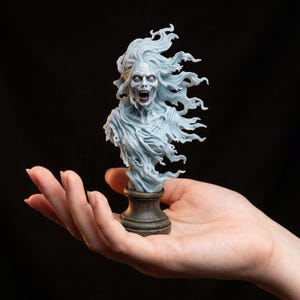 Op de afbeelding: Een bleke, licht blauwgrijze harsbuste van een schreeuwend, spookachtig figuur met golvend, warrig haar en flarden van etherische kleding. Het gedetailleerde beeldje is gemonteerd op een donkere, ronde voet en oogt zeer dynamisch.