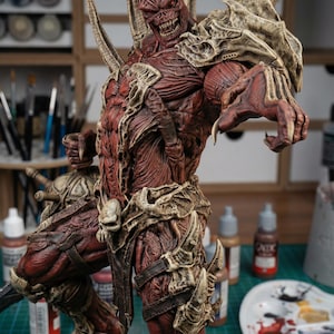 Può includere: Figura fantasy dettagliata, rossa e color osso. Presenta corna, decorazioni a teschio e un'armatura elaborata. La statua è posizionata su una base di teschi, mostrando un alto livello di artigianalità e dettaglio.