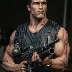 May include: A detailed statue of a man holding a rifle, wearing a black vest and a black shirt. The statue has a serious expression and is muscular. A grenade is attached to the vest. The statue is a collectible item.