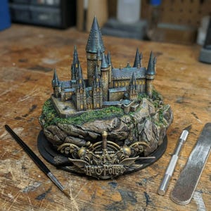 May include: A detailed miniature of Hogwarts Castle, featuring multiple towers and intricate architectural details. The base includes the words "Harry Potter" in a decorative emblem. The castle is set on a rocky landscape with green foliage.