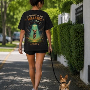 Puede incluir: Una camiseta negra con un estampado gráfico de un perro pequeño siendo abducido por un OVNI. El texto sobre el OVNI dice HE THOUGHT IT WAS WALKIES, y el texto debajo dice TINY BARK STUDIO. El diseño incluye un haz de luz verde brillante.