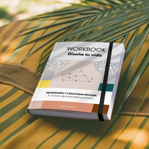 May include: A workbook titled "WORKBOOK Diseña tu vida" with a geometric design on the cover. The book is bound with a black elastic band and features text in Spanish. The background is a mix of yellow and green.