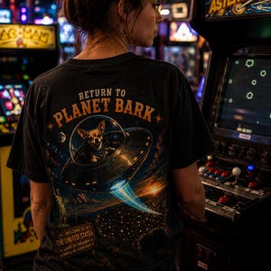 Puede incluir: Una camiseta negra con un estampado gráfico en la espalda. El diseño muestra un chihuahua en un platillo volante con el texto "RETURN TO PLANET BARK" encima. Debajo, un cartel dice "WELCOME TO THE UNITED STATES, LAND OF FREEDOM, HOME OF GOOD BOYS."