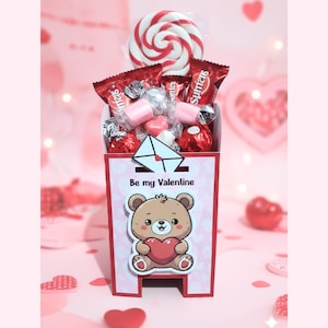 Puede incluir: Una caja de regalo de San Valentín con un oso de dibujos animados sosteniendo un corazón y el texto "Be my Valentine". La caja está llena de caramelos, chocolates y una piruleta roja y blanca.