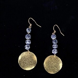 May include: Dangle earrings with gold-coloured hooks. Each earring has blue and white patterned beads, leading to a large, textured, gold-coloured disc. The earrings hang against a black background.