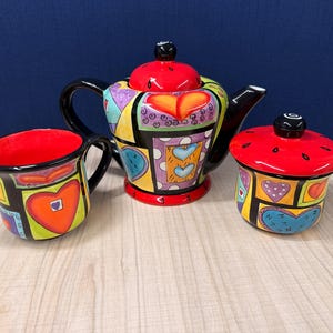 Könnte beinhalten: Ein Keramik-Teeservice bestehend aus einer Teekanne, einem Milchkännchen und einer Zuckerdose mit Deckel. Jedes Teil zeigt ein lebhaftes, mehrfarbiges geometrisches Muster mit Herzmotiven in Rot, Orange, Blau und Lila, schwarz umrandet.