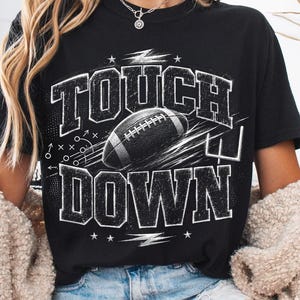 Könnte beinhalten: Ein schwarzes Kurzarm-T-Shirt mit einem verwaschenen weißen Grafikdruck, der den Text TOUCH DOWN, eine Football-Illustration und Spielzüge zeigt. Das Design umfasst zudem eine Torstangen-Grafik und kleine Sternakzente.