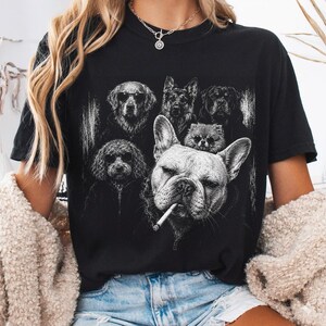 May include: A black short-sleeved t-shirt featuring a white graphic print of several dog breeds, including a French Bulldog with a cigarette in its mouth, a Golden Retriever, a German Shepherd, a Rottweiler, a Poodle, and a small Pomeranian.
