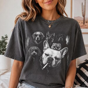May include: A charcoal gray short-sleeve t-shirt featuring a white graphic print of several dog breeds. A prominent French Bulldog with a cigarette in its mouth is centered, surrounded by smaller illustrations of other dogs, some wearing sunglasses.
