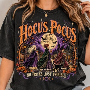 May include: A charcoal gray t-shirt featuring a graphic print of three figures in cloaks walking through a town at night under a full moon. The text HOCUS POCUS appears at the top in orange, with NO TRICKS, JUST TROUBLE printed at the bottom.