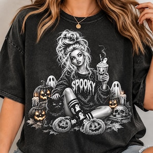 May include: A charcoal grey t-shirt featuring a graphic illustration of a person with a messy bun wearing a jumper with the word SPOOKY printed on it. The design includes a black cat, jack-o'-lanterns, ghosts, a skull, and a drink cup.