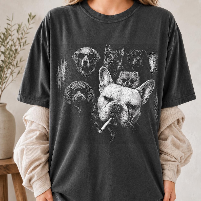 May include: A charcoal gray short-sleeve t-shirt featuring a white graphic print of several dogs. The central image shows a French Bulldog with a cigarette in its mouth, surrounded by other dog breeds, some wearing sunglasses, in a sketch style.