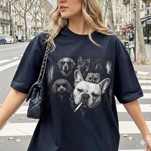 May include: A black short-sleeved t-shirt featuring a graphic print of several dog breeds, including a prominent French Bulldog with a cigarette in its mouth. The shirt is paired with a black quilted shoulder bag with a gold chain strap.