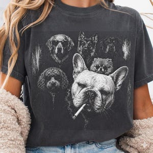 May include: A charcoal gray short-sleeve t-shirt featuring a white graphic print of five dogs. A French bulldog with a cigarette in its mouth is in the foreground, while four other dogs, including a poodle and a golden retriever, are behind it.