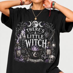 May include: A black t-shirt featuring a white graphic print of a cauldron, potion bottles, a skull, and candles. The text reads, "THERE'S A LITTLE WITCH IN ALL OF US," surrounded by moon phases, spiderwebs, and bare tree branches in a detailed illustration.
