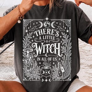 May include: A charcoal gray short-sleeve t-shirt featuring a white graphic print. The design includes the text "THERE'S A LITTLE WITCH IN ALL OF US" surrounded by illustrations of mushrooms, crystals, a cauldron, a broom, bats, and moon phases.